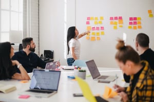 Lady standing at a white board with sticky notes talking and 4 co-workers around a table with computers collaborating with her.