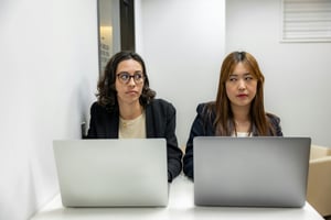Two ladies sitting next to each other in a close office space and looking at each other with contempt